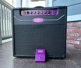 Budda guitar amplifier, black tolex, black grill cloth purple control panel with purple footswitch, sitting on wood floor, with brick wall behind it