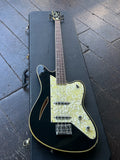 A black Charvel bass with a cream-colored pickguard and gold trim, resting inside an open black guitar case on a dark wooden deck floor.