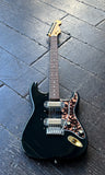 A black electric guitar with a tortoiseshell pickguard and gold hardware, resting on a dark wooden plank floor.