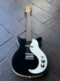 A black Danelectro twelve string electric guitar with a white pickguard and gold-toned hardware, photographed from above on a dark wood plank floor.