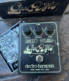 Electro-Harmonix God Wills modulator guitar pedal with black knobs and silver metal footswitch on a dark textured surface, with the branded box and patterned insert partially visible behind it.