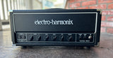 Electro-Harmonix MIG-50 guitar amplifier head in black with white lettering, photographed outdoors on a wooden surface with a brick wall and railing in the background.
