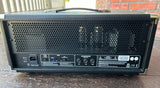 A black guitar amplifier head photographed outdoors on a wooden surface, showing the rear panel with input jacks, power cord, and control labels.