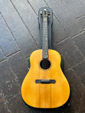 A natural wood acoustic guitar with an ebony body and neck, resting on a dark wooden plank floor.