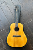 A Custom acoustic guitar with a natural wood body, ebony fret board and black headstock, resting diagonally on a black guitar case over a dark wooden floor.