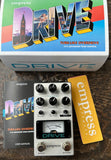 An Empress Drive tube-like overdrive guitar pedal shown with its retail box, user manual, and a yellow branded accessory on a dark wooden surface.