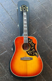 2025 Epiphone Hummingbird  acoustic guitar with red sunburst finish, featuring ornate gold floral inlays on a black pickguard, resting on a dark wooden floor next to its black hard case.