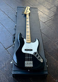 AFender  black electric bass guitar with a maple neck and white pickguard, resting on top of its black hard case on a dark wooden floor.