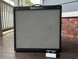 A Fender DeVille guitar amplifier cabinet standing on a wooden porch floor, with a small handwritten note placed in front of it. The background shows a brick wall, house siding, and a railing.