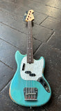 A Fender Mustang bass guitar in turquoise in a distressed finish, resting on a dark wooden plank floor. The headstock is visible at the top, showing the red and white logo of a Fender bass model.