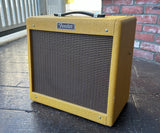A Fender Pro Junior guitar amplifier with  yellow tweed tweed covering and a dark brown front grille, photographed outdoors on a dark tiled floor with a brick wall in the background.