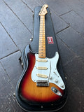 A Fender Stratocaster electric guitar in a sunburst finish, displayed diagonally inside its black hard case on a dark wooden plank floor.