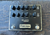 A Friedman BEzOD Deluxe guitar effects pedal with black knobs, silver footswitches, and a cream-colored border on a dark textured stone surface.