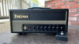 A Friedman guitar amplifier head with a black textured cabinet, gold lettering, and a front control panel, photographed on a wooden surface with a brick wall in the background.