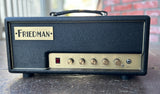A Friedman guitar amplifier head with a black textured leather cover and a cream-colored front control panel, resting on a dark wooden surface with a brick wall in the background.