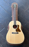 A Gibson J-35 acoustic guitar with a natural wood finish and a tiger-eye style pickguard, displayed vertically on a dark wooden plank floor.
