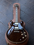 A Gibson ES335 dark brown glossy electric guitar with gold hardware, resting inside a brown leather hard case on a dark wooden floor.