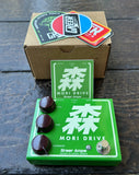 A green Mori Drive guitar pedal by Greear Amps with three dark knobs and a silver footswitch, placed on a dark wooden surface in front of its cardboard box and stickers.