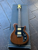 A vintage Gretsch solid body electric guitar , chrome hardware, two humbucker tan pick ups, with a brown wood body and black pickguard, resting on top of its black hard case on a dark wooden deck floor.