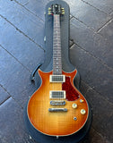 A High Spirit double-cutaway electric guitar with a sunburst finish, resting inside a black hard case on a dark wooden plank floor. The guitar features a flamed maple top, a red glittery pickguard, gold hardware, and a black headstock with gold lettering.