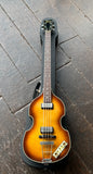 Hofner Violin bass in sunburst with chrome pick ups, rosewood fretboard, black headstock resting on case displayed on wooden floor