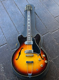 A sunburst Gibson ES-330TD electric guitar with a black body and neck, resting on a dark wooden plank floor. The guitar features gold hardware and chrome pickups.