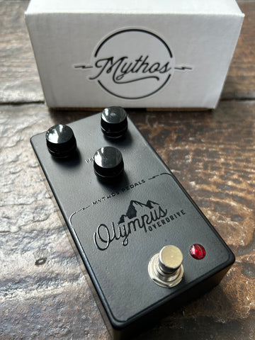 Black effect pedal with 'Olympus Overdrive' branding on a wooden surface, with a white box labeled 'Mythos' in the background.
