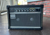 A Roland Jazz Chorus-25 guitar amplifier sitting on a concrete patio in front of a brick wall. The amplifier is black with a silver grille and features the Roland logo on the left side.