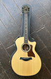 A Taylor acoustic-electric guitar with a natural wood body and dark pickguard, resting on a dark gray textured surface.