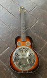 A Dobro vintage resonator guitar with a sunburst finish, featuring a large silver metal resonator dome on the body, resting on a dark wooden plank floor.