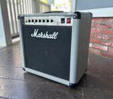 A Marshall guitar amplifier with a gray textured grille and black front speaker grille, sitting on a dark wooden deck with a brick wall in the background.