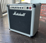 A grey Marshall guitar amplifier with a gray textured cabinet, black front grille, and white Marshall logo, photographed outdoors on a wooden deck with a brick wall in the background.