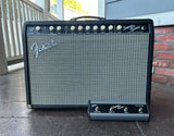 A black Fender Super Sonic guitar amplifier head with a matching foot switch, photographed outdoors on a patio against a brick wall and railing.