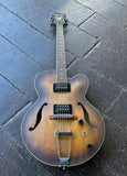 Ibanez hollow body a double-cutaway electric guitar with a natural sunburst tobacco finish, resting on a dark gray stone-tiled floor. The guitar features chrome hardware, a chrome tailpiece, and a headstock with a visible logo.