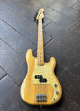 An Ibanez natural wood electric bass guitar with a glittering yellow pearloid pickguard and black hardware, photographed from above on a dark wooden floor.