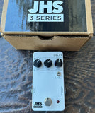 A JHS 3 Series Delay guitar effects pedal with black knobs and a silver footswitch on a white faceplate, shown in front of its brown cardboard box on a dark textured surface.