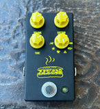 A JHS black guitar effects pedal with four yellow knobs labeled Volume, Tone, Sustain, and an additional top knob, featuring a yellow hot dog illustration and a silver foot switch on a textured dark surface.