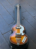 A Jay Turser Violin Bass with a sunburst finish, silver hardware, and a white pickguard, resting inside a black hard case on a dark tiled floor.