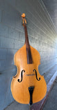 A wooden Kay double bass instrument leaning against a gray concrete block wall in a covered passage.
