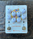 A white Caverns by Keeley guitar effects pedal with multiple white control knobs, red text labels, and gold-toned footswitches, photographed from above on a dark textured surface.