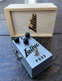 A Lollar Fuzz guitar effects pedal with a silver metal casing, black knobs, and a silver footswitch, shown in front of its light wood carrying case on a dark wooden tabletop.