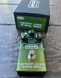 An MXR Carbon Copy Analog Delay guitar pedal in metallic green with black knobs and a silver footswitch, shown in front of its black retail box on a dark wooden surface.