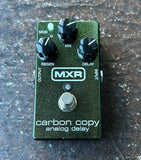 An MXR Carbon Copy analog delay guitar effects pedal with a textured dark green metallic body, four black control knobs, and a silver footswitch, resting on a dark textured stone surface.