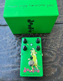 A green Doobie pedal guitar effect unit with black knobs and a yellow dog illustration, placed in front of a matching green box on a dark wooden surface.