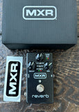 An MXR Reverb guitar effects pedal shown with its black MXR box and a white sticker on a dark wood surface. The pedal is black with silver controls and a silver footswitch.
