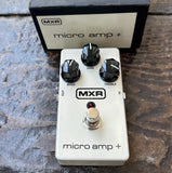 MXR Micro Amp + guitar effects pedal in white with black knobs and a silver footswitch, shown with its black and white box on a textured stone surface.