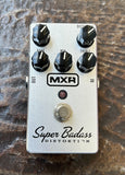 MXR Super Badass Distortion guitar effects pedal with a silver metal body, black knobs, and a silver footswitch, photographed on a textured brown surface.