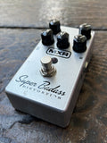 MXH silver Super Badass Distortion guitar pedal with a brushed silver metal body, black knobs, and a silver footswitch, resting on a dark wooden surface.