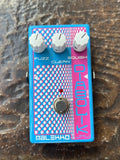 A Malekko FX-CW-00D electric guitar effects pedal with a blue and pink faceplate, three white knobs, and a silver footswitch, resting on a textured brown surface.