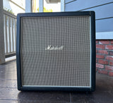 A Marshall speaker cabinet standing on a porch, with a black frame, silver mesh grille, and the Marshall logo centered on the front.